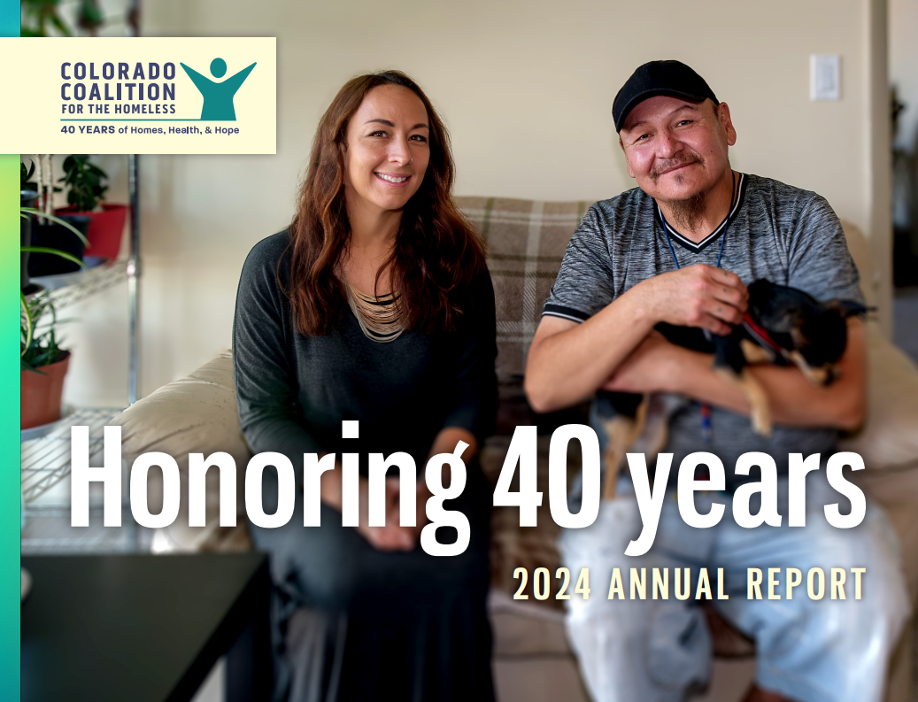 two individuals sit on a couch facing the camera. Person on right holds a small dog. 